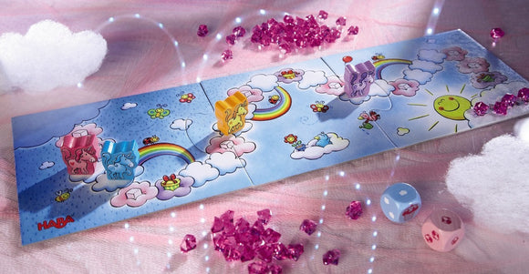 A vibrant game setup featuring Unicorn Glitterluck Cloud Crystals by Haba. The game board displays colorful illustrations of rainbows, clouds, and flowers, with playful unicorn figures in various colors positioned on the board. Sparkling pink crystal-like shapes add a whimsical touch, while two colorful dice are placed beside the game, enhancing the magical atmosphere.