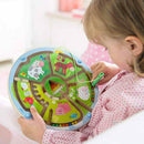 A child engages with the HABA Magnetic Game Number Maze. The colorful circular maze features farm animal illustrations and numbered sections, with a green string tool being used to guide a bead through the maze. The child is seated, wearing a patterned shirt, focused on the game that promotes fine motor skills and number recognition.