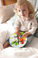 A young child playing with the HABA Motor Skills Game. The circular wooden puzzle features colorful pieces that fit into various slots, promoting fine motor skill development. The child looks focused and engaged while seated comfortably with pillows around them.