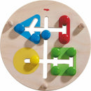 A colorful mechanical motor skills game by HABA featuring a circular wooden base with various shaped cut-outs and vibrant wooden pegs in red, blue, yellow, and green. Kids can manipulate the pegs through the shapes, enhancing their fine motor skills and coordination.