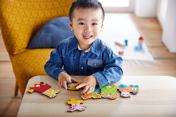 A cheerful young child focuses on assembling a colorful jigsaw puzzle featuring a number train, with various animal-shaped pieces displayed on a wooden table. The child is dressed in a denim shirt and is sitting in a bright room, suggesting an engaging playtime experience.