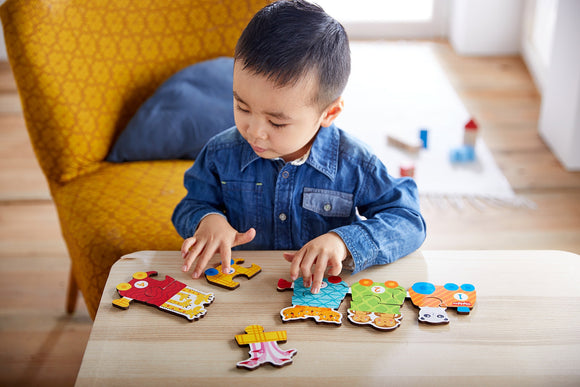 A young child wearing a denim shirt is focused on assembling colorful jigsaw pieces on a wooden table. The vibrant puzzle features animal motifs and numbers, promoting early learning and fine motor skills. The cozy setting includes a yellow patterned chair in the background.