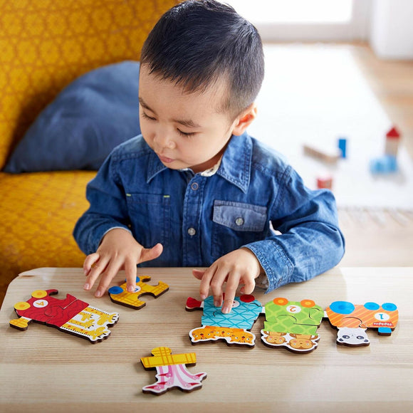 A young child is assembling the HABA Number Train jigsaw puzzle on a wooden table. The puzzle pieces are colorful and display numbers and playful designs, including animals. A yellow patterned sofa can be seen in the background.