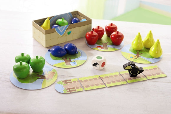 My First Orchard game by HABA USA featuring colorful fruit pieces, including green apples, red apples, blue plums, and yellow pears. The game includes a wooden die and a black crow figure. The components are arranged on circular game boards depicting trees and fruit paths, encouraging interactive play for young children.