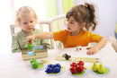 Two young children engaged in playing 'My First Orchard' by HABA USA. One child, wearing an orange shirt, reaches towards a box filled with colorful fruit pieces, while the other child, in a green striped shirt, observes with focus. The game components include 3D fruit figures, cards, and a die, arranged on a bright white tabletop in a well-lit room.