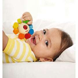 A happy baby lying on a soft surface, holding a colorful Clutching Toy Whirlygig by Haba. The toy features bright, circular segments and a wooden base, perfect for developing fine motor skills.