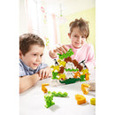 Two children playing the Animal Upon Animal game from HABA. One child, a boy, is concentrated as he places green and colorful animal pieces on top of a wooden animal tower while the girl, wearing a pink sweater, smiles and gestures as she prepares to add her piece. The background is bright and playful, enhancing the fun, engaging atmosphere of the game.