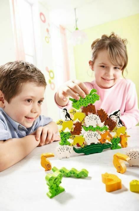 Two children playing with the HABA Animal Upon Animal game, featuring a colorful stacking arrangement of various animal figures including sheep, penguins, and lizards. The kids are focused on building the animal tower on the table in a bright, cheerful environment.
