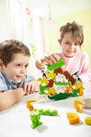 Two children playing with the HABA Animal Upon Animal game, featuring a colorful stacking arrangement of various animal figures including sheep, penguins, and lizards. The kids are focused on building the animal tower on the table in a bright, cheerful environment.