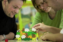 Three adults engaging in a fun game of 'Animal Upon Animal' by HABA. They are balancing colorful wooden animal pieces on a tabletop. The scene shows smiles and excitement as players try to stack their animals without toppling them over. A red die is placed on the table near them, adding to the game's playful atmosphere.