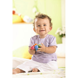 A joyful baby sitting on a soft surface, holding colorful clutching toys. The baby has short curly hair and is wearing a light purple shirt with a white skirt. Bright, playful surroundings include soft toys and greenery, creating a cheerful atmosphere for play.