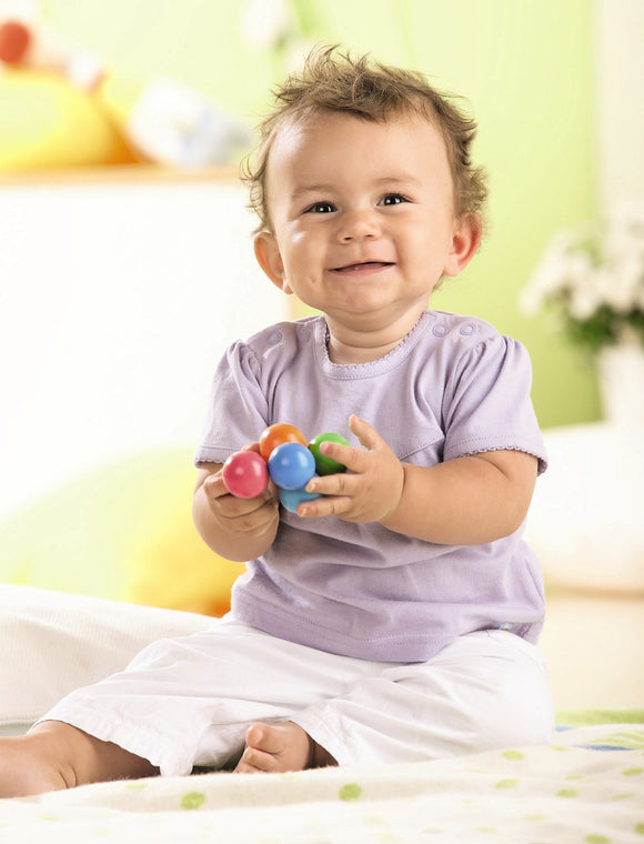 A cheerful baby sitting on a soft surface, playing with colorful clutching toys. The baby has a joyful expression and is holding vibrant plastic balls in various colors, promoting sensory play and motor skill development.