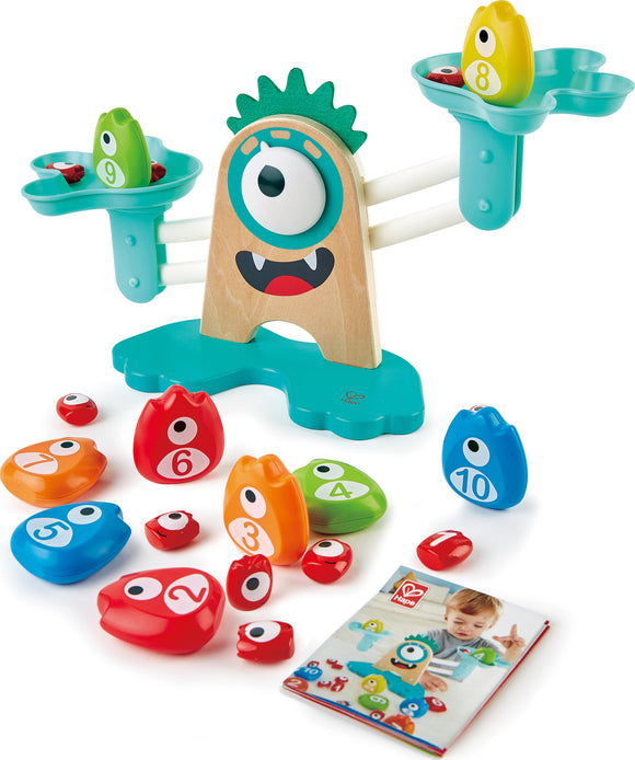 The Monster Math Scale by HaPe International features a fun, colorful design with a wooden base shaped like a monster. It includes two balancing scales and various numbered monster shapes in vibrant colors, each labeled with a number. The set comes with a booklet showing a child engaging with the scale, promoting early math skills through a playful experience.