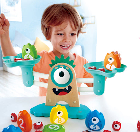 A young boy joyfully plays with the Monster Math Scale by HaPe International. The playful scale features colorful monster-shaped weights and is designed to engage children in learning basic math concepts while having fun. The boy smiles as he balances the colorful weights on either side of the scale, enhancing his cognitive skills through interactive play.