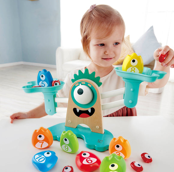 A young child engages with the Monster Math Scale toy by HaPe International, featuring a colorful, monster-themed design with number blocks. The child is holding a red number block, while other colorful blocks are arranged on a scale, which displays a whimsical monster face.
