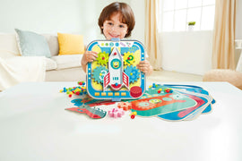 A cheerful child holds the Cogworks Experimental Playset by Hape, featuring a colorful rocket design with interlocking gears. Surrounding the child are various colorful pieces and activity boards, all placed on a bright, uncluttered table. The background showcases a cozy living space.