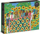The Sunflower Quilting Bee at Arles jigsaw puzzle by Galison features a vibrant illustration of diverse figures gathering around a quilt with sunflowers, set against a colorful background of greenery and blue sky. The puzzle comes in a sleek black box showcasing the artwork.