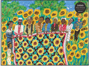 The Sunflower Quilting Bee at Arles jigsaw puzzle by Galison featuring a vibrant illustration of a group of diverse women, gathered around a large quilt adorned with colorful sunflowers, set against a bright blue background dotted with sunflowers. The scene captures a sense of community and artistry, ideal for puzzle enthusiasts.