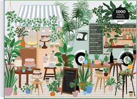 A colorful and intricately illustrated jigsaw puzzle titled 'Plant Cafe' by Galison. The scene depicts a vibrant outdoor cafe filled with various plants, including succulents, palms, and ferns. Beautiful desserts are displayed on wooden tables, enhancing the inviting atmosphere. A charming food truck is visible in the background, adorned with signage detailing plants and cafe offerings. The puzzle consists of 1000 pieces, perfect for plant lovers and puzzle enthusiasts alike.