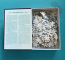 Open box of the jigsaw puzzle 'Murder Most Puzzling: The Missing Will' by Chronicle Books. The box includes a partially filled bag of white jigsaw pieces and a page of text detailing a mysterious storyline involving inheritance and a detective investigation.
