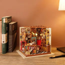 A detailed view of Sam's Study Model Kit by Robotime, showcasing a miniature study scene. The kit features shelves filled with books, a cozy red chair, delicate lights illuminating the space, and a ladder, all crafted with intricate attention to detail. Nearby are two open books resting on a wooden table, enhancing the inviting ambiance of the study.