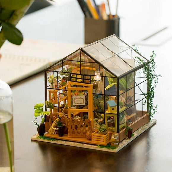A detailed model kit of Cathy's Flower House by Robotime, showcasing a miniature greenhouse filled with vibrant plants, a yellow facade, and charming garden accessories. The clear roof and glass walls allow a peek into the intricately designed interior, perfect for DIY enthusiasts.