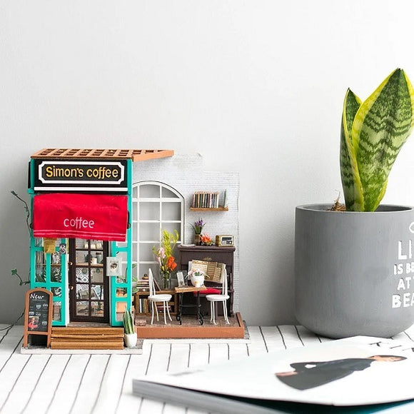 Simon's Coffee Model Kit featuring a detailed miniature coffee shop with a sign reading 'Simon's coffee,' a red awning, a window with books, and a cozy interior setup with a table and chairs. Accompanied by a potted plant and an open magazine for a homely atmosphere.