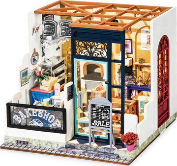 A detailed DIY miniature model of Nancy's Bake Shop by Robotime, featuring a cozy storefront with an intricate sign, a chalkboard advertisement outside, and a glimpse of the warm interior full of bakery items and décor.