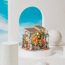 A beautifully detailed DIY model kit of Emily's Flower Shop, showcasing a small, charming building with a front display of potted plants, flowers, and a cozy interior with warm lighting. The structure includes intricate design elements like a sign, windows, and decorative accents that create an inviting atmosphere.