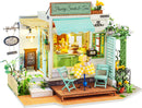 A detailed model kit of a charming tea and sweets shop, featuring an inviting exterior with pale green walls, a bright blue awning, and a wooden deck. The storefront displays an array of treats inside, and there are two mint green chairs at a yellow checkered table on the patio. The scene is adorned with decorative flowers and a welcoming sign, capturing a cozy café atmosphere perfect for DIY enthusiasts.