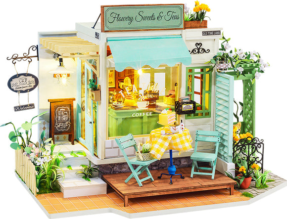 A detailed model kit of a charming tea and sweets shop, featuring an inviting exterior with pale green walls, a bright blue awning, and a wooden deck. The storefront displays an array of treats inside, and there are two mint green chairs at a yellow checkered table on the patio. The scene is adorned with decorative flowers and a welcoming sign, capturing a cozy café atmosphere perfect for DIY enthusiasts.