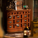 Kiki's Magic Emporium by Robotime is a detailed DIY kit featuring a beautifully crafted wooden chest of drawers. The scene includes labeled drawers with names like 'Dittany' and 'Unwound,' a decorative key, and a sparkling gem, all set in a warm, inviting atmosphere. Perfect for enthusiasts of miniature craftsmanship.