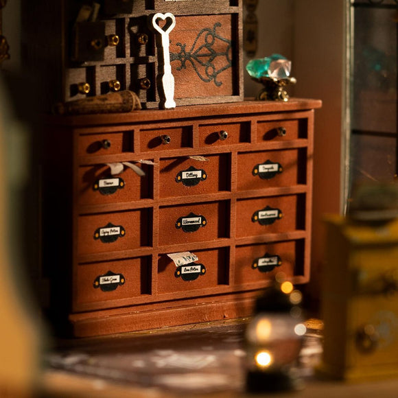 Kiki's Magic Emporium by Robotime is a detailed DIY kit featuring a beautifully crafted wooden chest of drawers. The scene includes labeled drawers with names like 'Dittany' and 'Unwound,' a decorative key, and a sparkling gem, all set in a warm, inviting atmosphere. Perfect for enthusiasts of miniature craftsmanship.