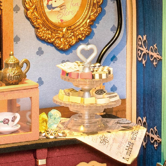 A charming display from Alice's Tea Store by Robotime, featuring a beautiful multi-tiered cake stand with various colorful sweets, a decorative teapot, and an elegant tea cup set. The background is adorned with floral patterns and a gold-framed picture that highlights the tea store theme.