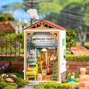 A detailed model kit depicting a charming fruit store named 'Morning Fruit'. The shop features a red rooftop, wooden frames, and a colorful display of fruits and vegetables. There's a welcoming sign, a ladder against the wall, and decorative plants, all set in a vibrant garden environment.