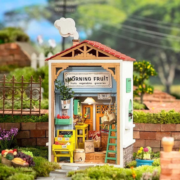 A detailed model kit depicting a charming fruit store named 'Morning Fruit'. The shop features a red rooftop, wooden frames, and a colorful display of fruits and vegetables. There's a welcoming sign, a ladder against the wall, and decorative plants, all set in a vibrant garden environment.