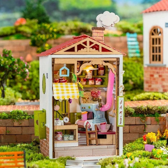 Sweet Jam Shop Model Kit showcasing a miniature shop interior. The wooden structure features a cozy design with shelves lined with colorful jars of jam, a green counter, and a pink machine. A yellow checkered tablecloth covers a small table, and soft light illuminates the space through a charming lamp. The scene is set in a lush green garden with brick pathways in the background.