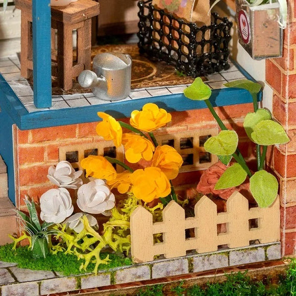 A detailed view of the Dream Yard Model Kit by Robotime, featuring a beautifully designed miniature scene with vibrant yellow flowers, white flowers, and decorative greenery, alongside a small wooden table and a watering can, all enclosed by a quaint fence.