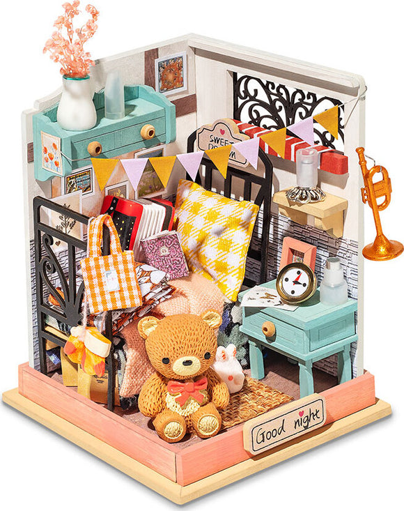 A detailed, colorful miniature model kit called Sweet Dreams Bedroom by Robotime. The kit features a cozy bedroom scene with items such as a teddy bear, decorative pillows, a small blue nightstand, a vase with flowers, and a miniature trumpet, all arranged in a charming corner setup. The walls are adorned with pictures and festive bunting, creating a warm and inviting atmosphere.
