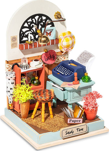 A detailed model kit by Robotime featuring a cozy study scene. The model includes a blue typewriter, a desk with a 'Paper' sign, colorful plants, and decorative items like a lamp and a hanging heart-shaped sign that says 'lucky'. The scene is richly decorated, encouraging creativity and providing a charming ambiance.