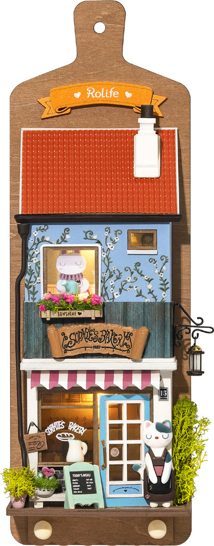 Aroma Toast Lab by Robotime is a charming DIY kit featuring a miniature bakery scene. The design showcases a cute blue building with a red roof, colorful flowers in window boxes, and a playful character outside. The bakery is adorned with details like a menu board, a lantern, and vibrant décor, inviting creativity and imagination.