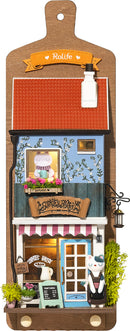 Aroma Toast Lab by Robotime is a charming DIY kit featuring a miniature bakery scene. The design showcases a cute blue building with a red roof, colorful flowers in window boxes, and a playful character outside. The bakery is adorned with details like a menu board, a lantern, and vibrant décor, inviting creativity and imagination.