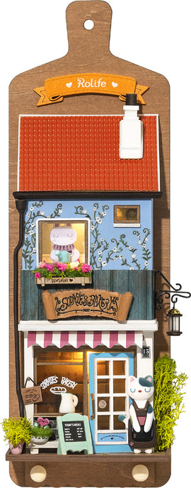 Aroma Toast Lab by Robotime is a charming DIY kit featuring a miniature bakery scene. The design showcases a cute blue building with a red roof, colorful flowers in window boxes, and a playful character outside. The bakery is adorned with details like a menu board, a lantern, and vibrant décor, inviting creativity and imagination.