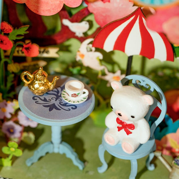 A whimsical DIY kit named Bear's Sweetie by Robotime, featuring a tiny white bear with a red bow sitting on a blue chair next to a decorative table. The table holds a golden teapot and a small cup adorned with a floral design, surrounded by vibrant paper flowers and greenery, creating a delightful miniature garden scene.