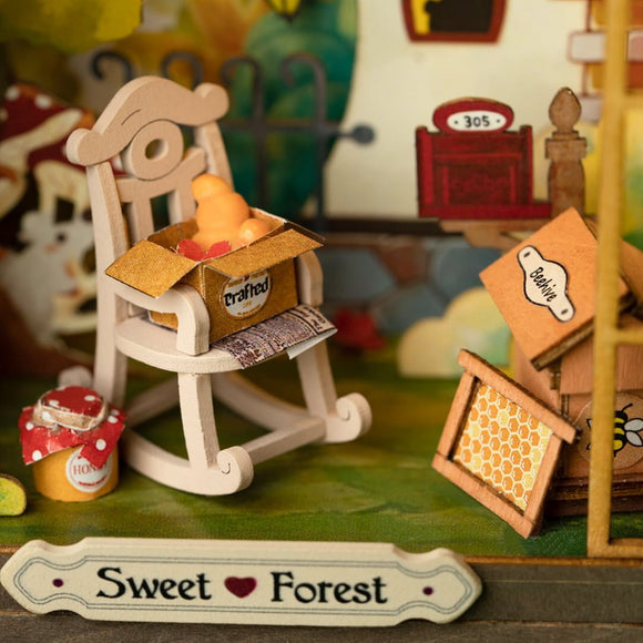The Sweet Forest DIY kit by Robotime showcases a charming miniature scene with a wooden rocking chair holding a crafted box filled with colorful objects, honey pots, and whimsical signs, set in a vibrant forest backdrop. Perfect for creative minds, this kit encourages imagination and hands-on crafting.
