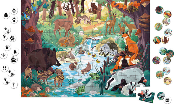 The Footprint Seek & Find Puzzle by Janod features a vibrant and colorful jigsaw scene depicting a lush forest with various animals including deer, foxes, and a boar near a stream, surrounded by trees and foliage. Accompanying the puzzle are circular tokens showcasing animal tracks, enhancing the interactive search experience for kids.