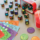 Fiesty Dice by MindWare displayed on a table, featuring colorful, fun dice with various expressive monster faces. The image shows multiple dice, game cards in vibrant colors, and tokens, all arranged around a red tin container. The game encourages creativity and quick thinking.