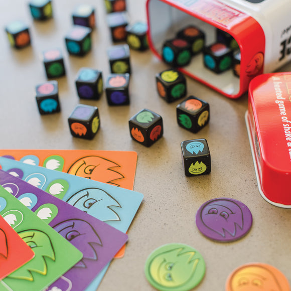 Fiesty Dice by MindWare displayed on a table, featuring colorful, fun dice with various expressive monster faces. The image shows multiple dice, game cards in vibrant colors, and tokens, all arranged around a red tin container. The game encourages creativity and quick thinking.