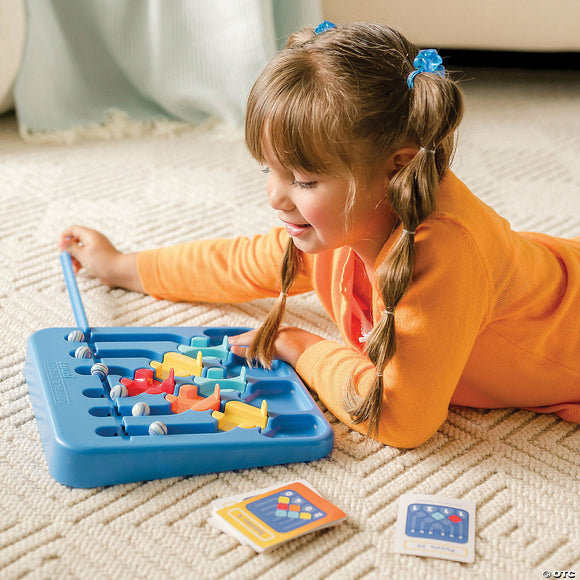 A young girl with pigtails engages with the Marble Circuit Jr. mechanical puzzle by MindWare. She is focused on guiding colorful marbles through a structured tray using a blue mechanism. The background features a cozy indoor setting, and cards with various challenges are placed on the white carpet beside her.