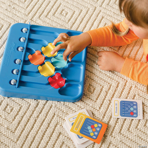 A child interacting with the Marble Circuit Jr. mechanical puzzle by MindWare, featuring a blue base with sliding colored puzzle pieces and solution cards nearby. The puzzle encourages critical thinking and fine motor skills.